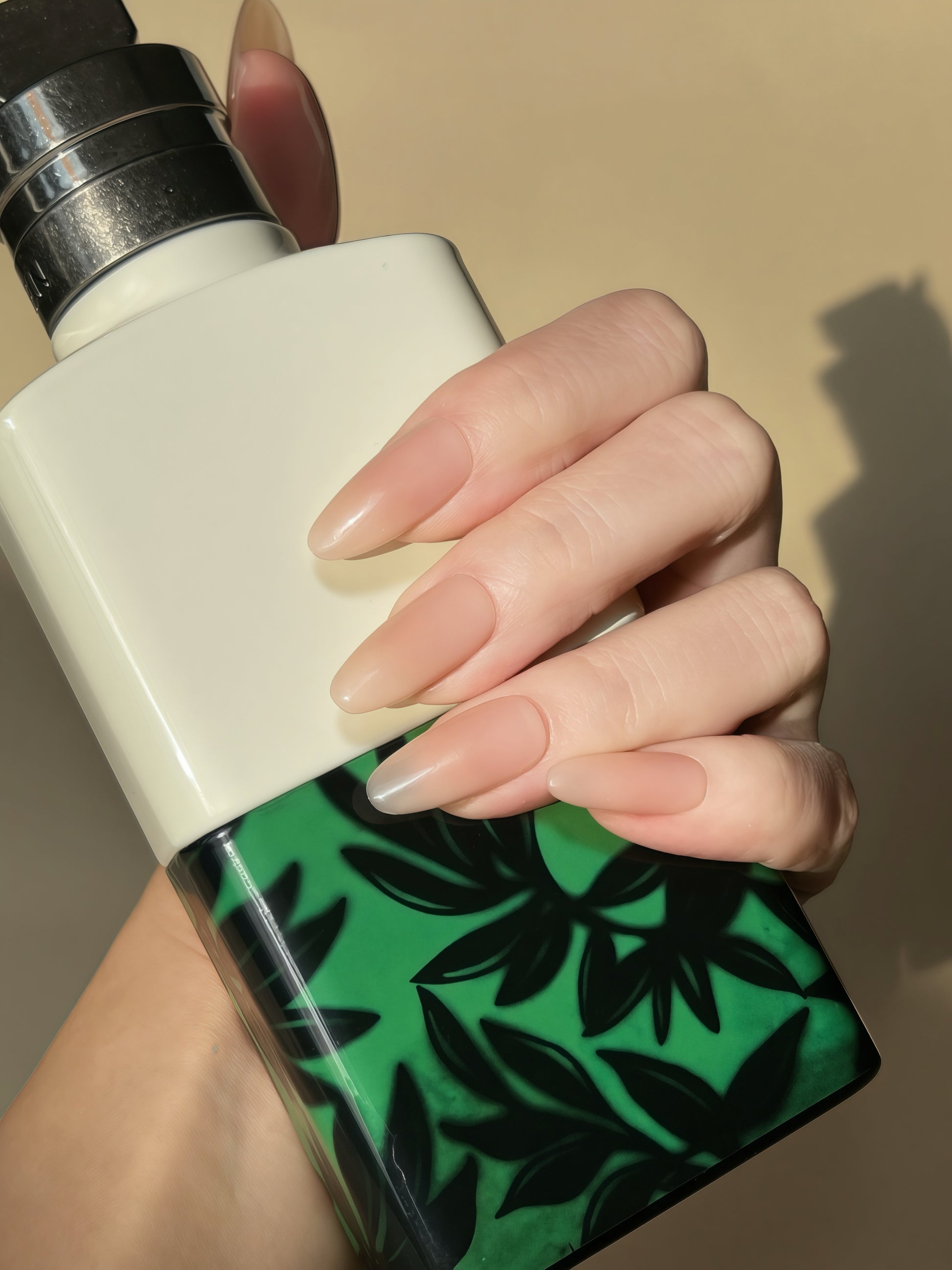 Hand wearing Bare Eternity nude oval press-on nails holding a green patterned bottle