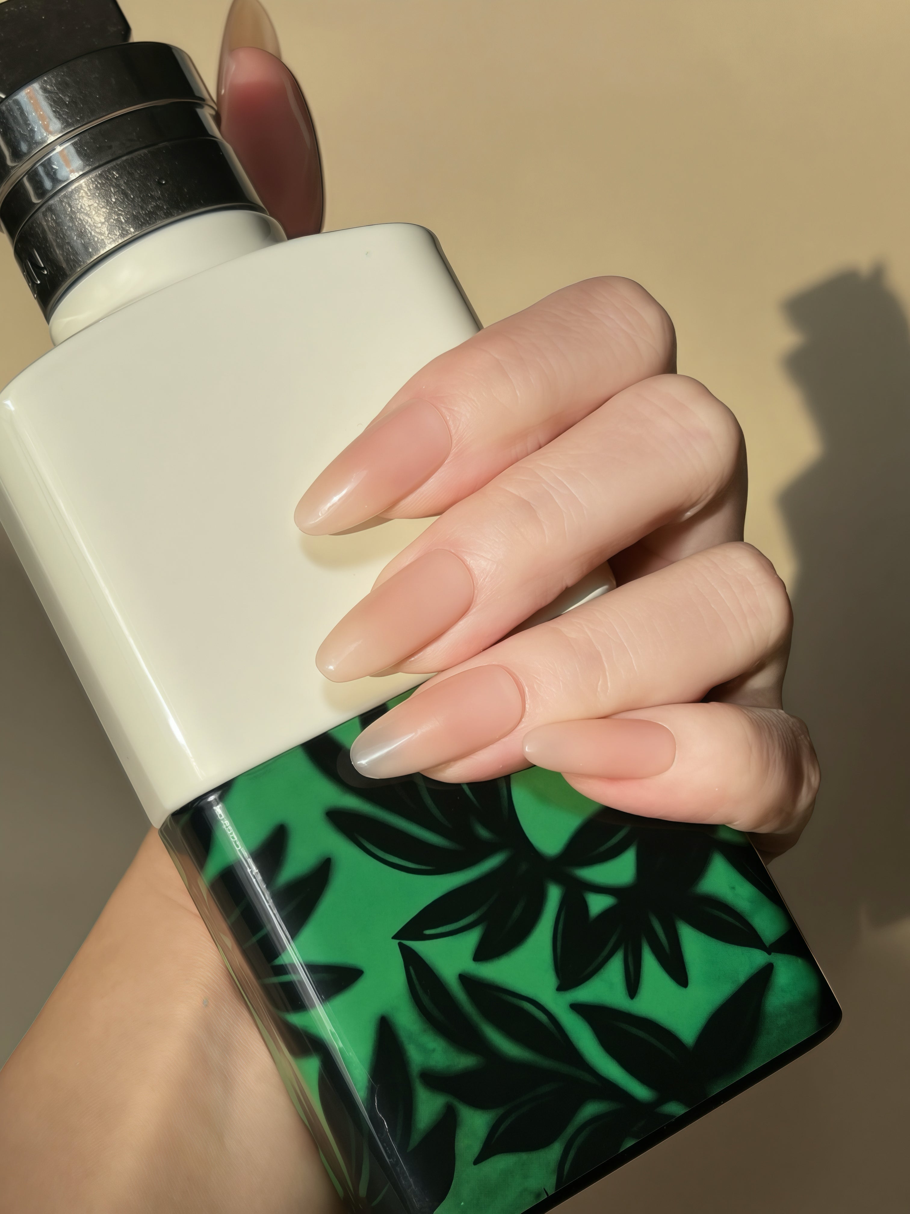 Hand wearing Bare Eternity nude oval press-on nails holding a green patterned bottle
