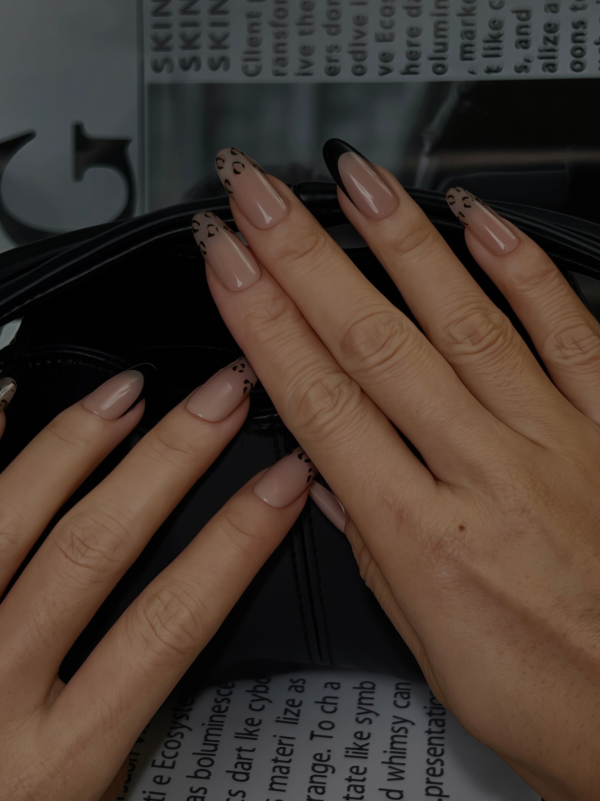 Long oval nude French press-on nails featuring subtle leopard print tips, displayed on both hands over a magazine background.