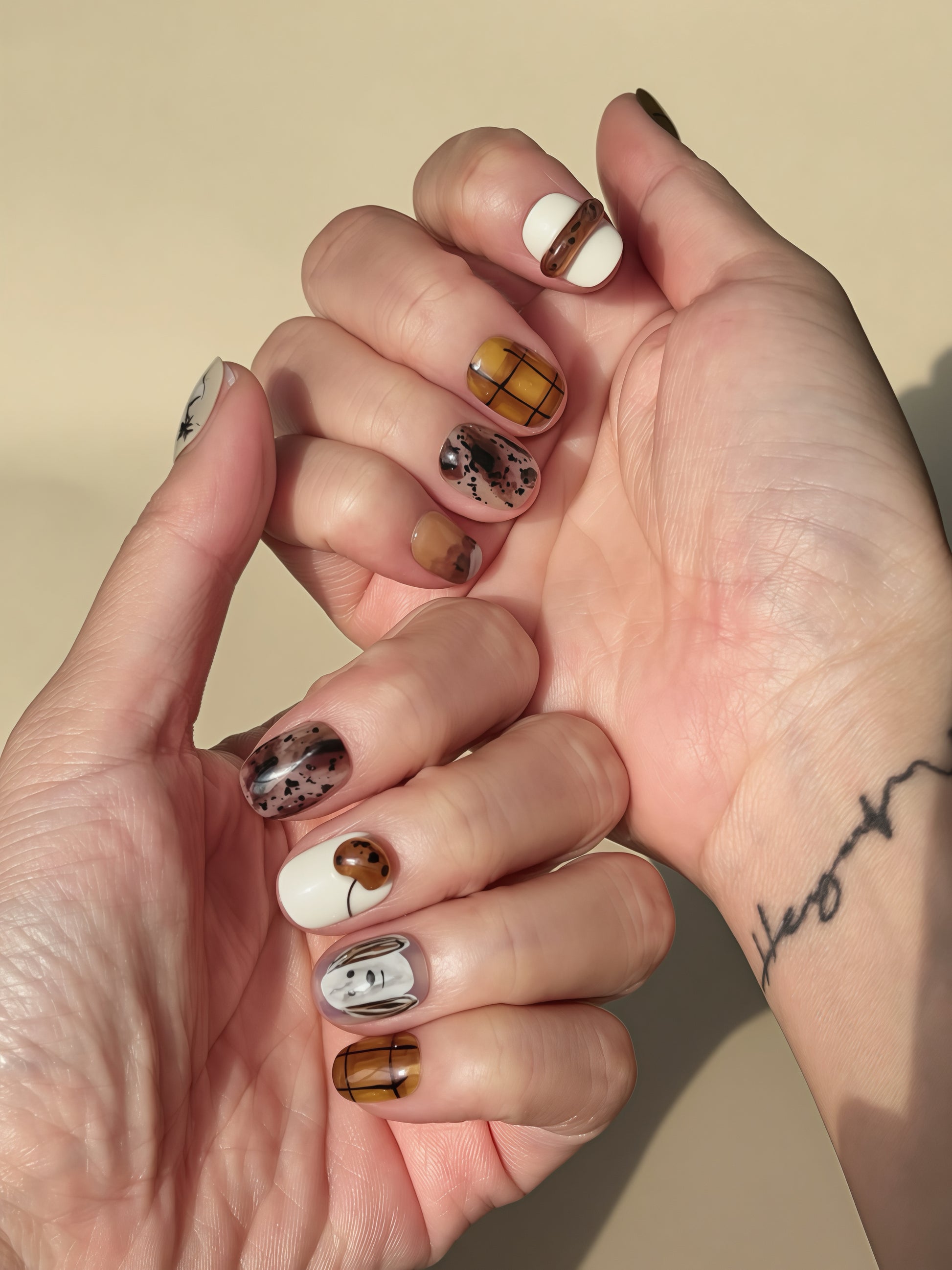 Warm brown and white press-on nails with hand-painted puppy doodles, coffee-tone plaid designs, and abstract speckled accents. Cozy autumn nail set shown on hands under natural sunlight.