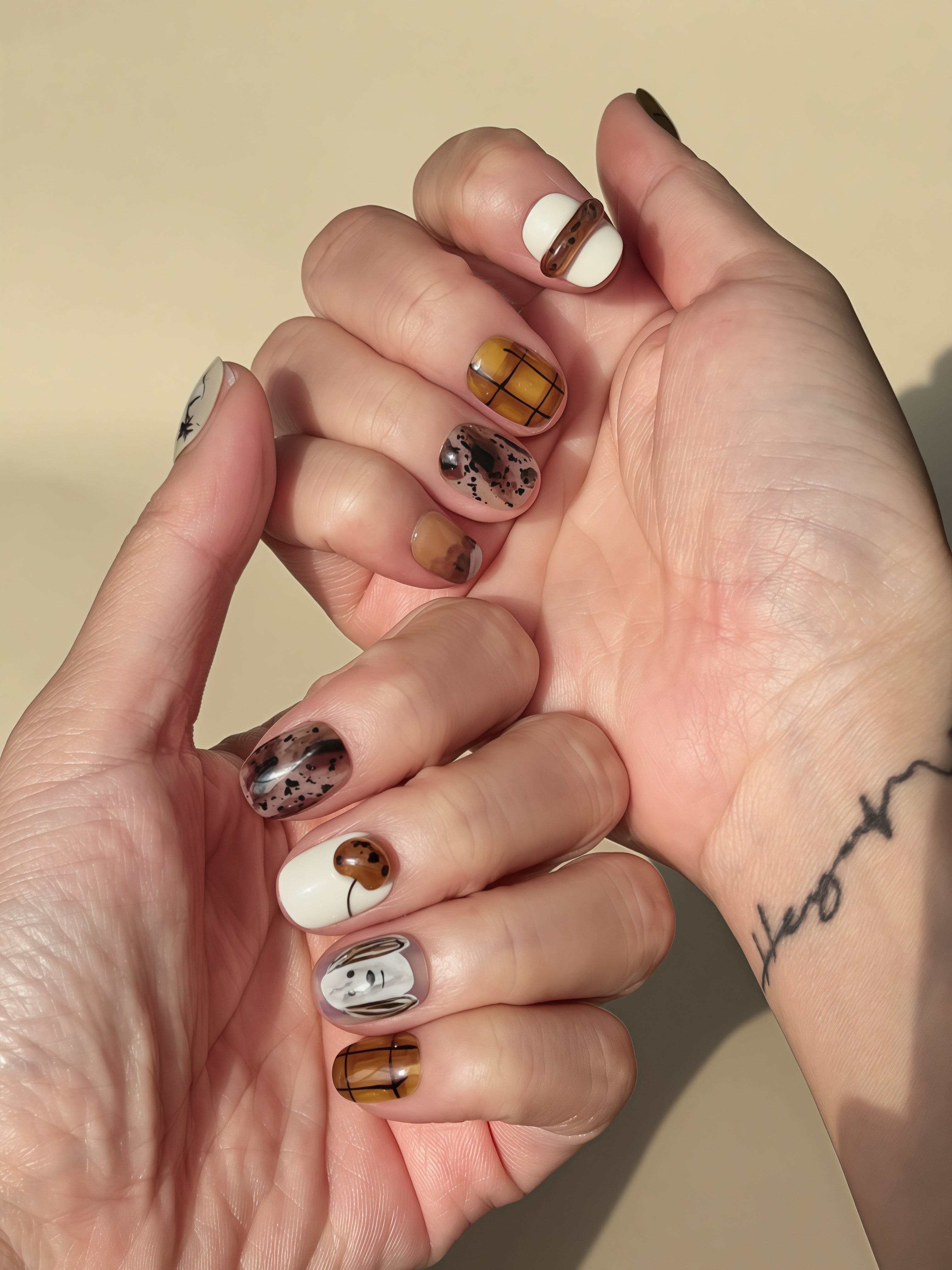 Warm brown and white press-on nails with hand-painted puppy doodles, coffee-tone plaid designs, and abstract speckled accents. Cozy autumn nail set shown on hands under natural sunlight.