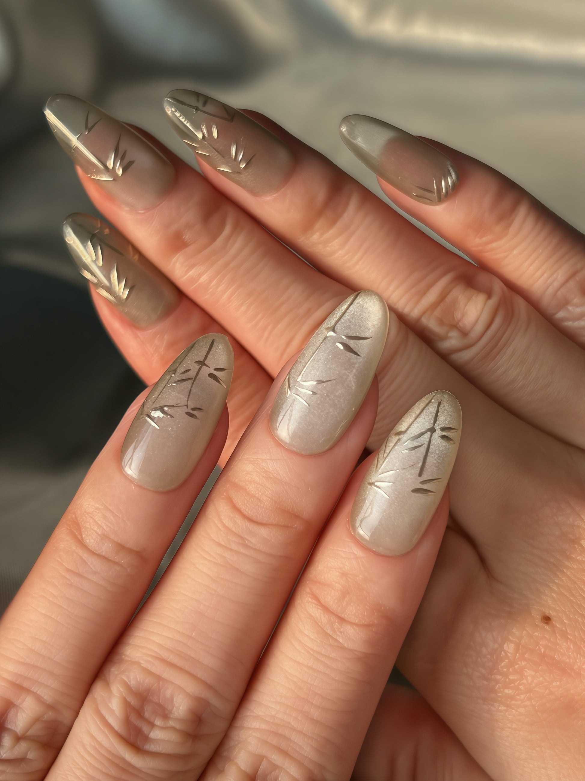 Shadow Bamboo nails on model hand in soft daylight — oval pearl-grey design with gold bamboo strokes.